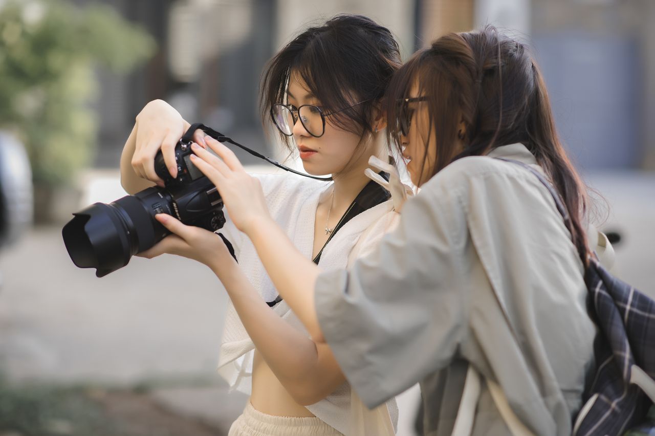 Offerings Two young women examining a DSLR camera outdoors, engaged in photography and learning.