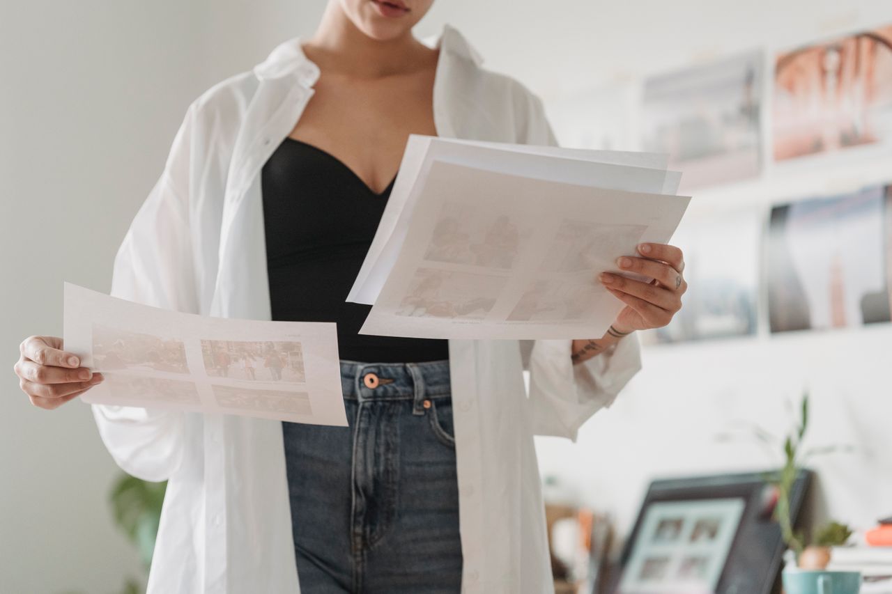 Offerings Crop anonymous female employee comparing photos on paper sheets while working on project in light house