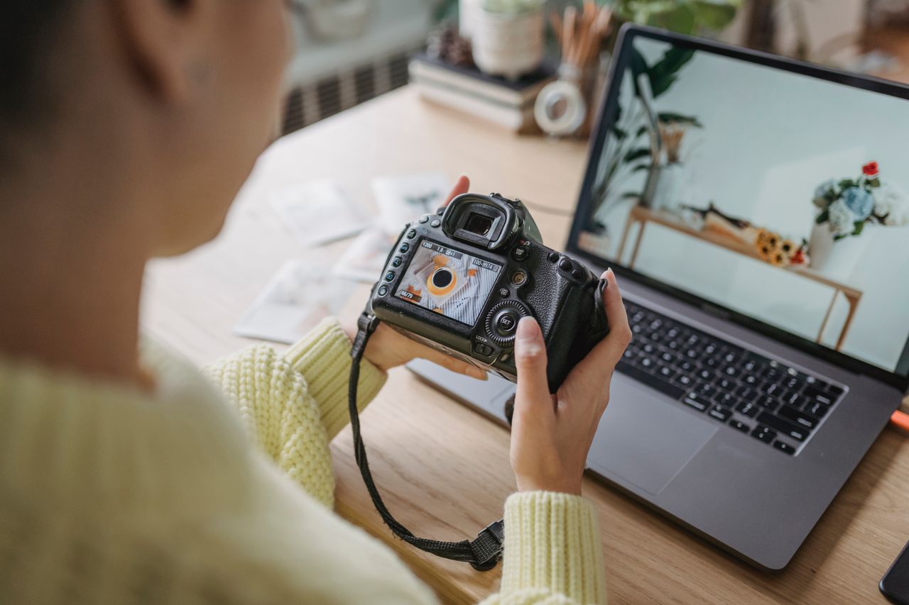 Offerings Unrecognizable crop female photographer looking at screen of photo camera while sitting at wooden table with netbook during work in light room